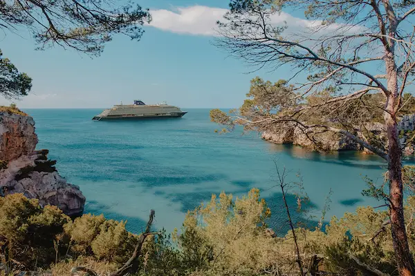 Cruisescapes luxury ocean cruise ship sailing near scenic pine-covered coastal cliffs in turquoise waters.